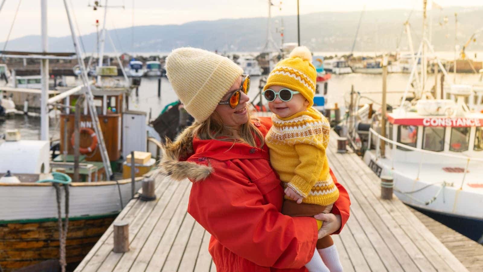 Mor kledd i rød jakke og hvit lue holder sitt barn kledd i gul genser og lue på en brygge med båter i bakgrunnen.